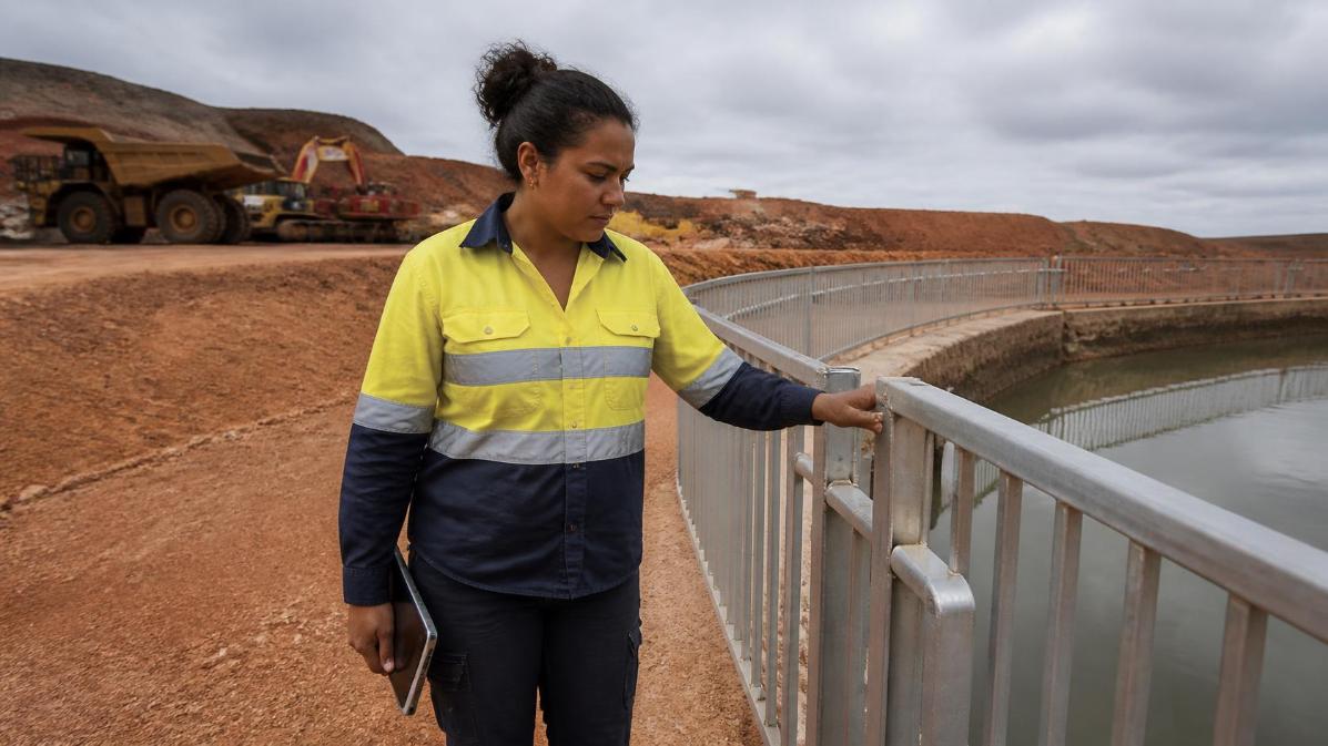 A worker inspects a steel water barrier fence beside a mining site retention pond.
